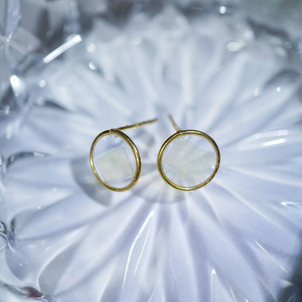 A pair of gold hoop earrings with a square-shaped white inlay, displayed on a textured, light-colored glass surface.
