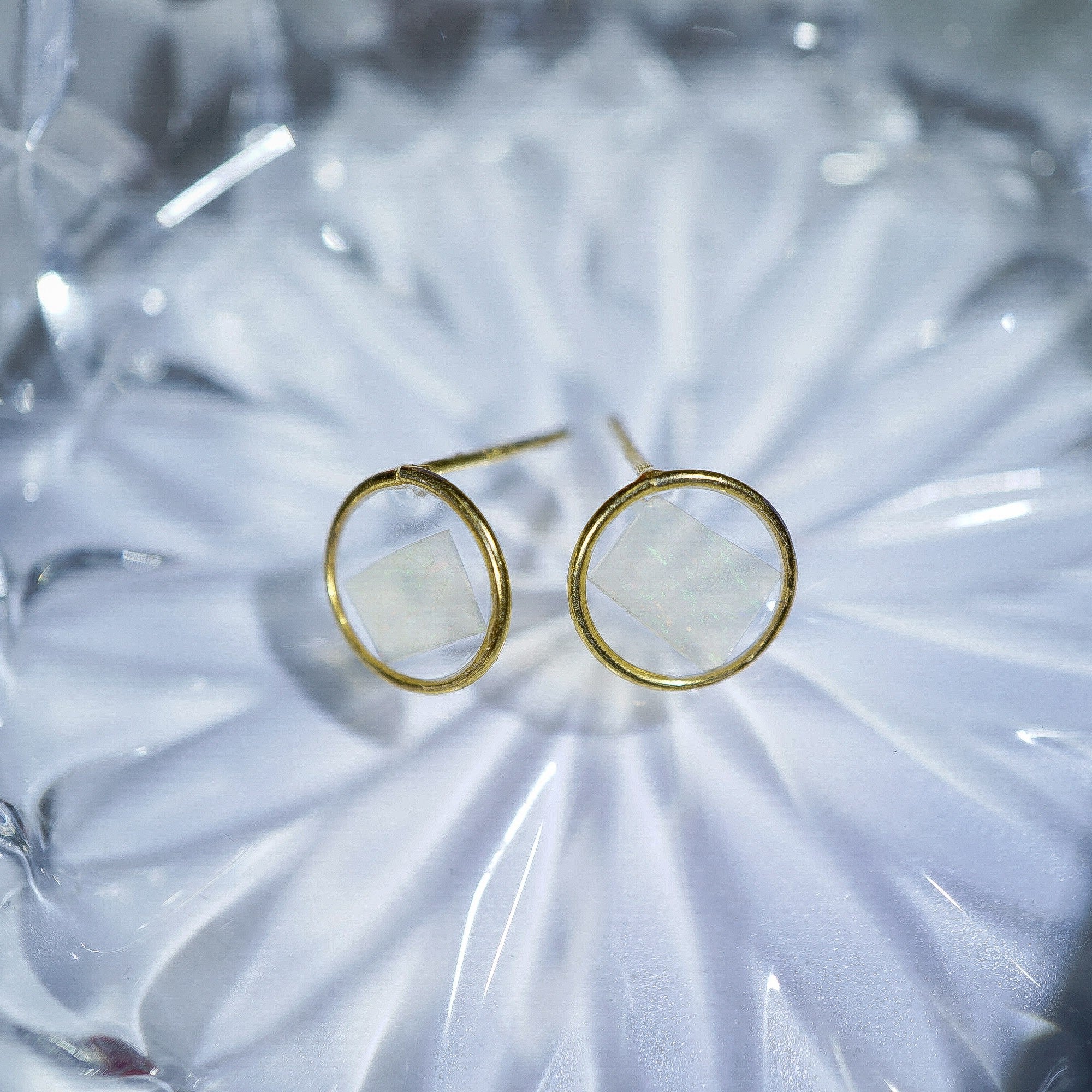 A pair of gold hoop earrings with a square-shaped white inlay, displayed on a textured, light-colored glass surface.
