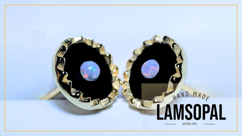 A pair of Lamsopal hand-made earrings featuring round black opal centers with a blue iridescent play-of-color, surrounded by a textured gold bezel setting. The image includes a 'HAND MADE LAMSOPAL' text overlay on the bottom right.