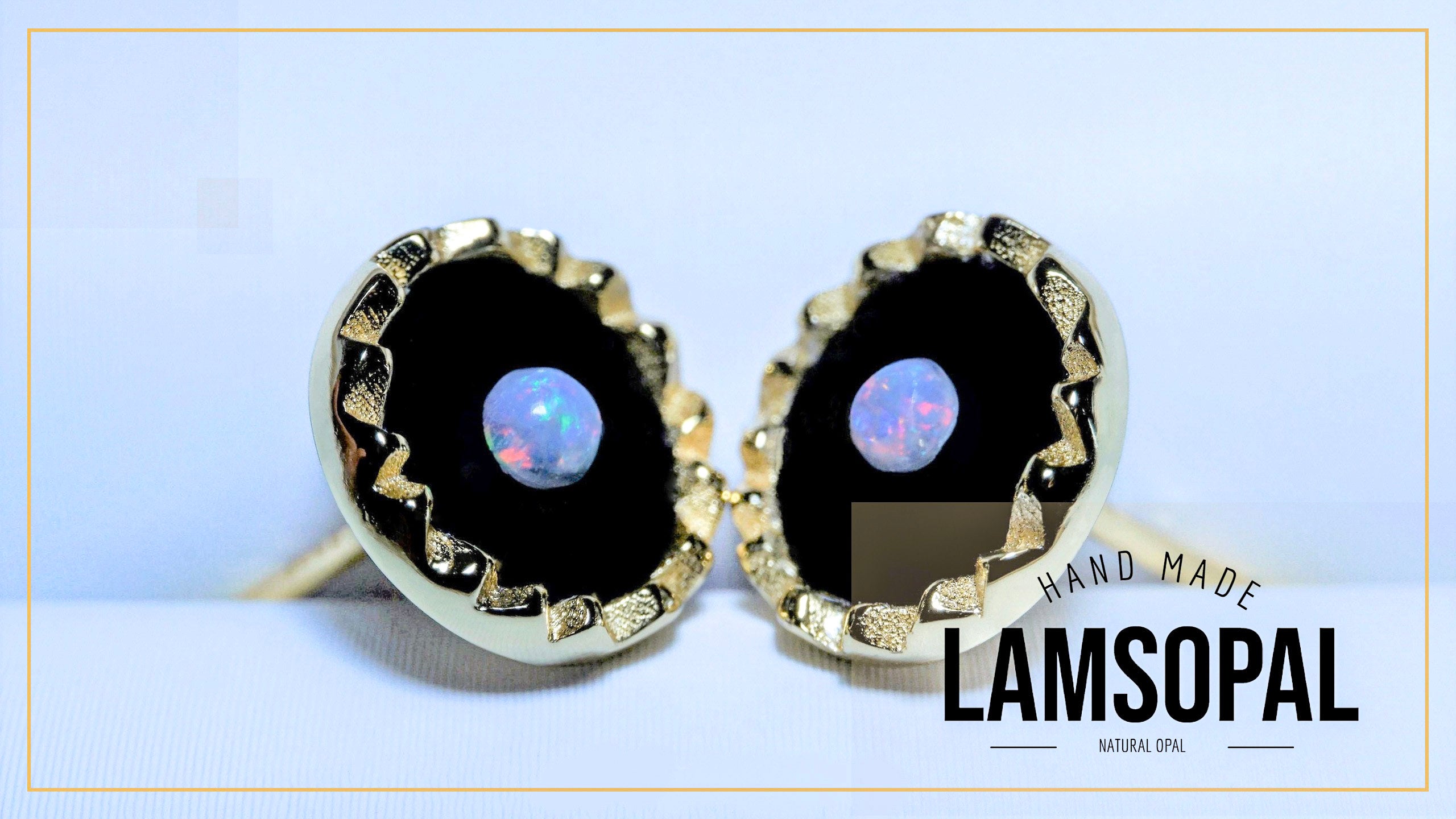 A pair of Lamsopal hand-made earrings featuring round black opal centers with a blue iridescent play-of-color, surrounded by a textured gold bezel setting. The image includes a 'HAND MADE LAMSOPAL' text overlay on the bottom right.