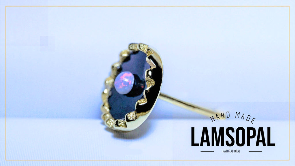 A handmade Lamsopal natural opal ring featuring a dark black opal inlay with a central pinkish-purple opal stone, surrounded by a gold-toned bezel with small clear crystals, shown on a light blue background with a brand logo overlay.