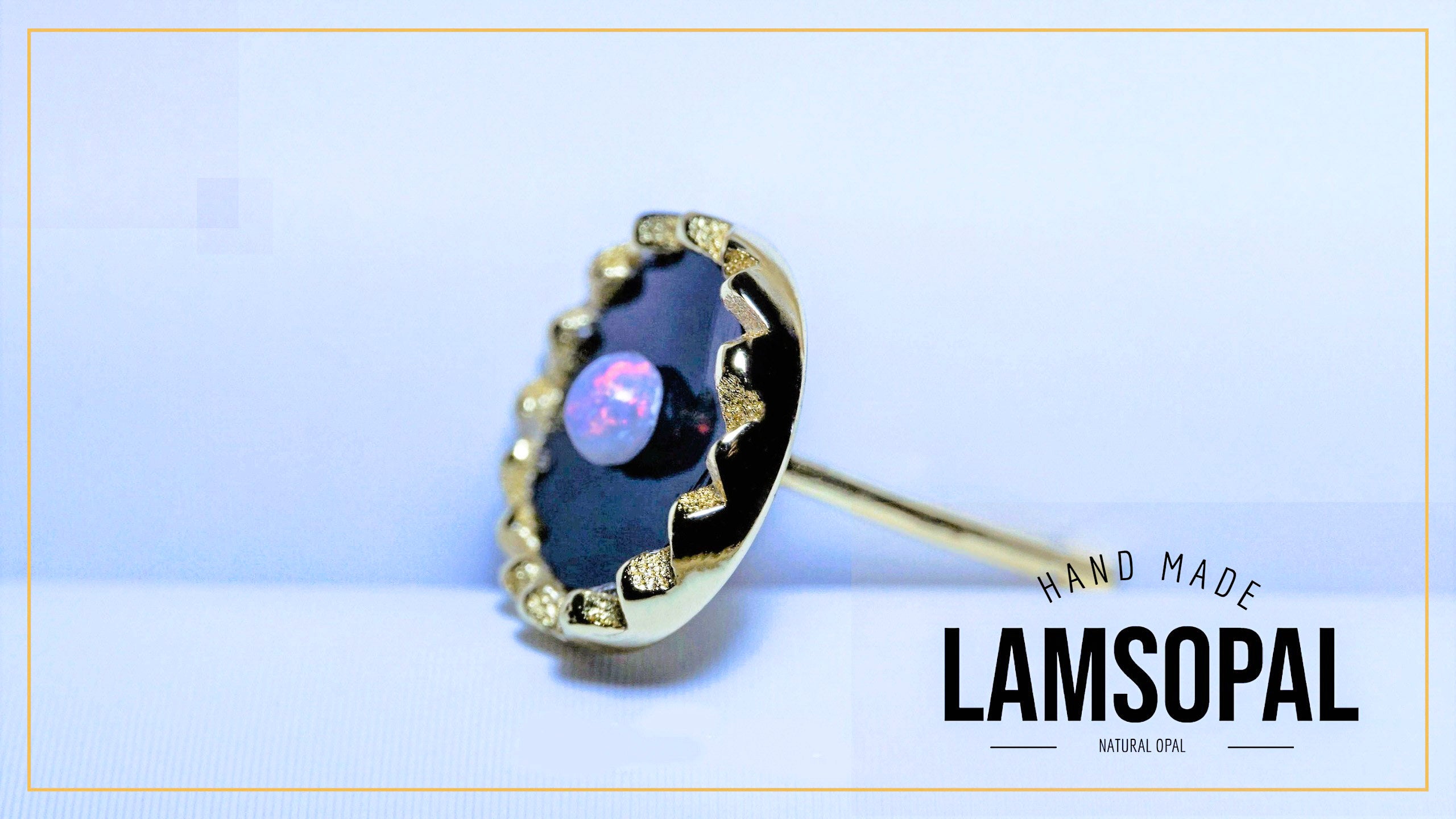 A handmade Lamsopal natural opal ring featuring a dark black opal inlay with a central pinkish-purple opal stone, surrounded by a gold-toned bezel with small clear crystals, shown on a light blue background with a brand logo overlay.