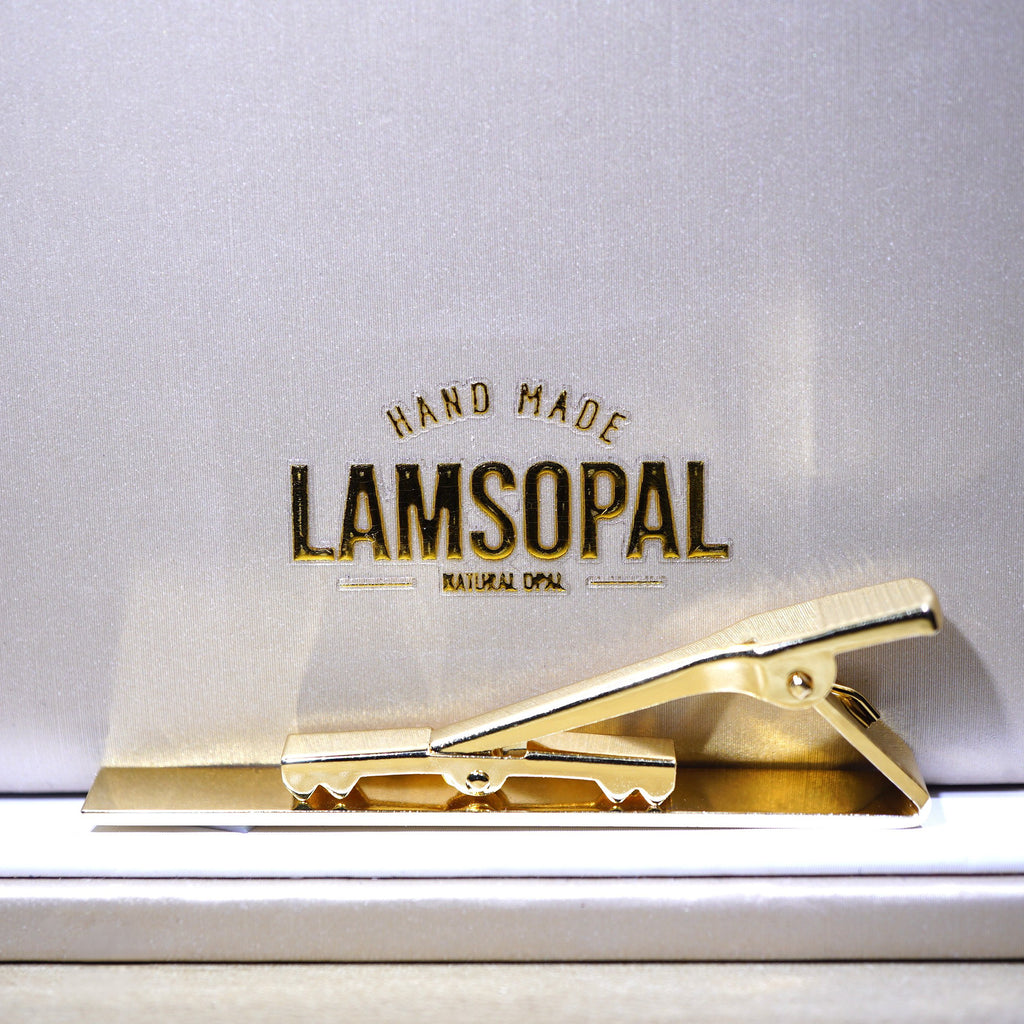 A gold metal tie clip resting on a white surface in front of a white background card printed with the brand name 'LAMSOPAL' and the text 'HAND MADE NATURAL COTTON'.