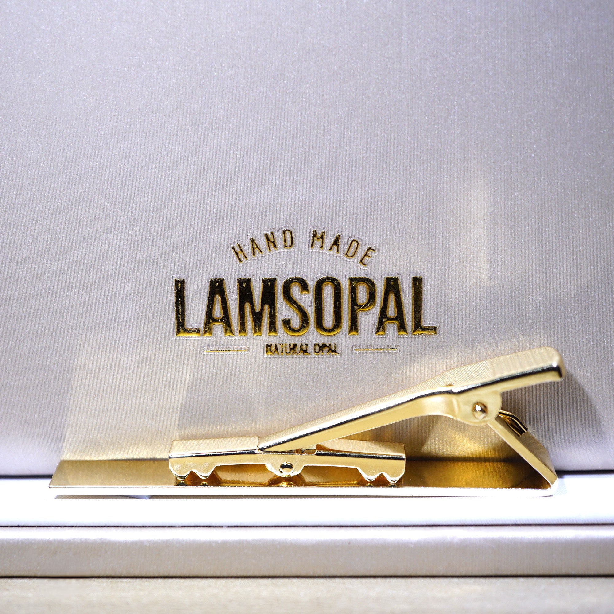 A gold metal tie clip resting on a white surface in front of a white background card printed with the brand name 'LAMSOPAL' and the text 'HAND MADE NATURAL COTTON'.