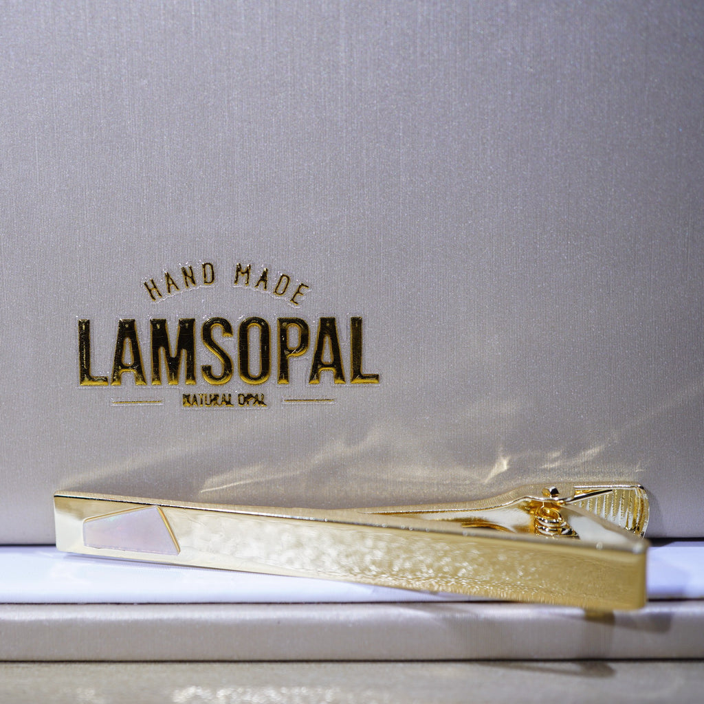 A gold tie bar with a textured pattern and a white inlay, resting on a white surface in front of its branded silver packaging box.