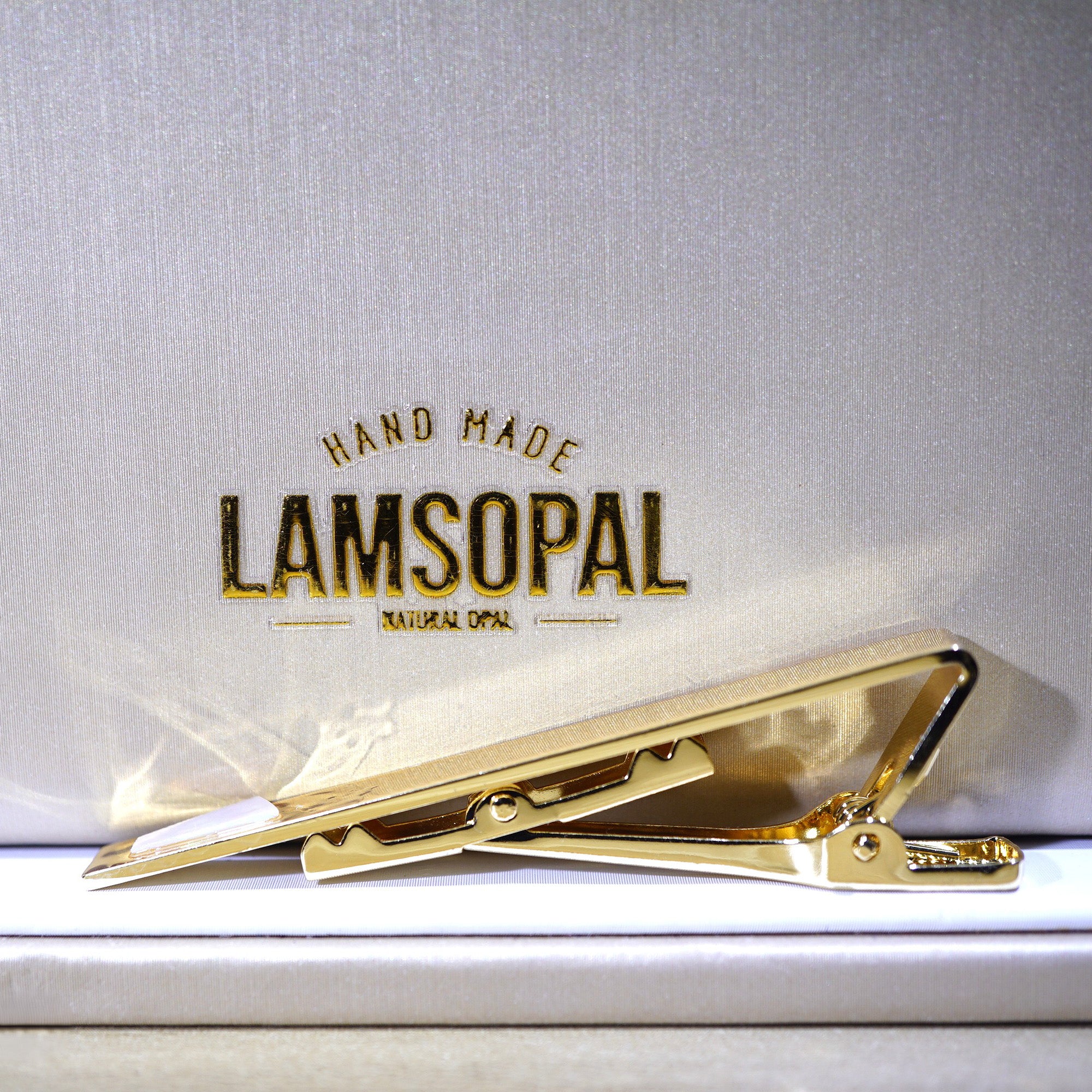 A gold tie clip displayed on a white textured background with the brand name 'LAMSOPAL' printed above it.