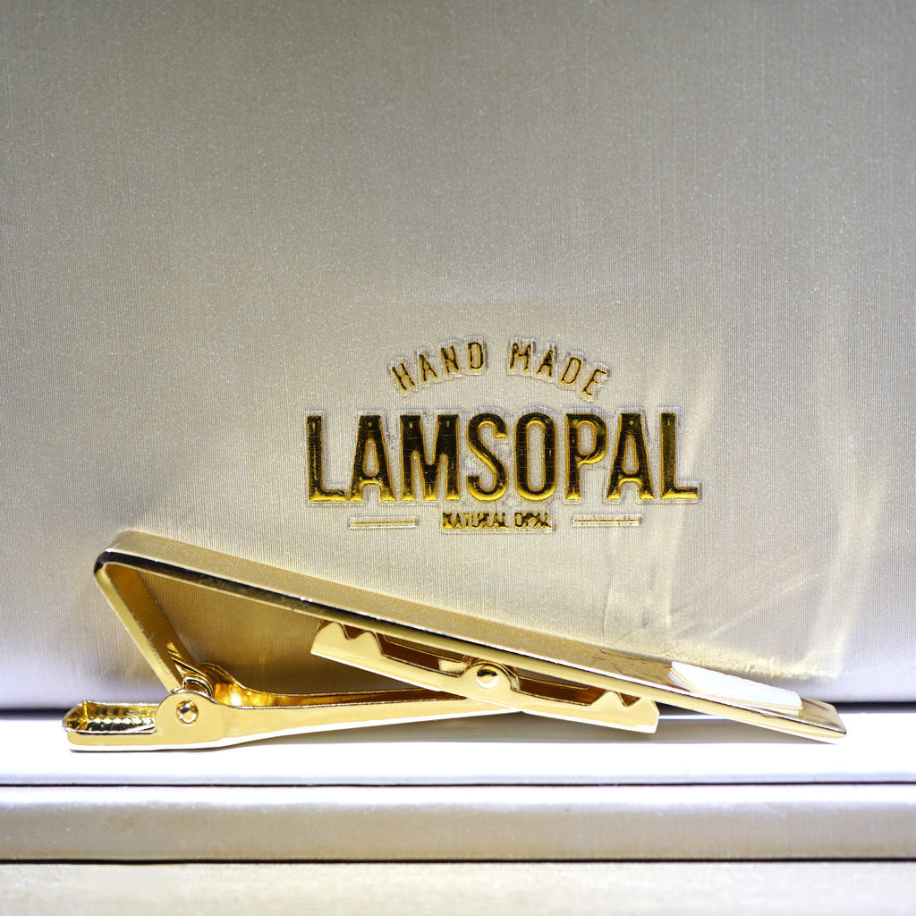 A gold tie bar clip resting on a white textured presentation box lid with the brand name 'LAMSOPAL' embossed in gold behind it.