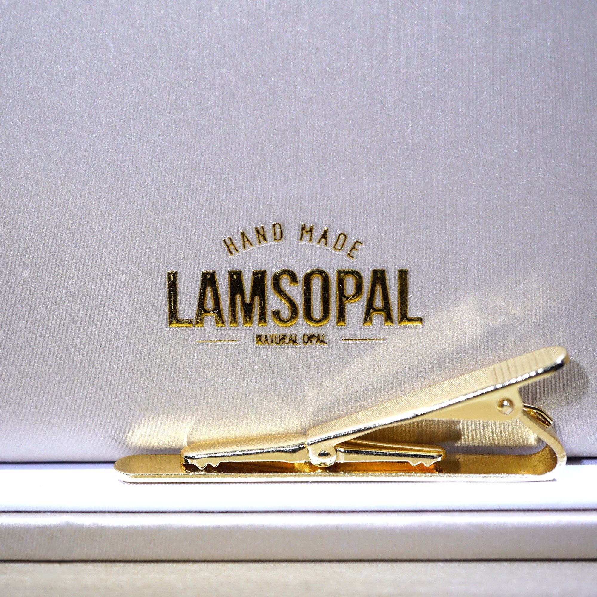 A gold metal hair clip resting on a white surface in front of a textured light gray box with the brand name 'LAMSOPAL' printed in gold.