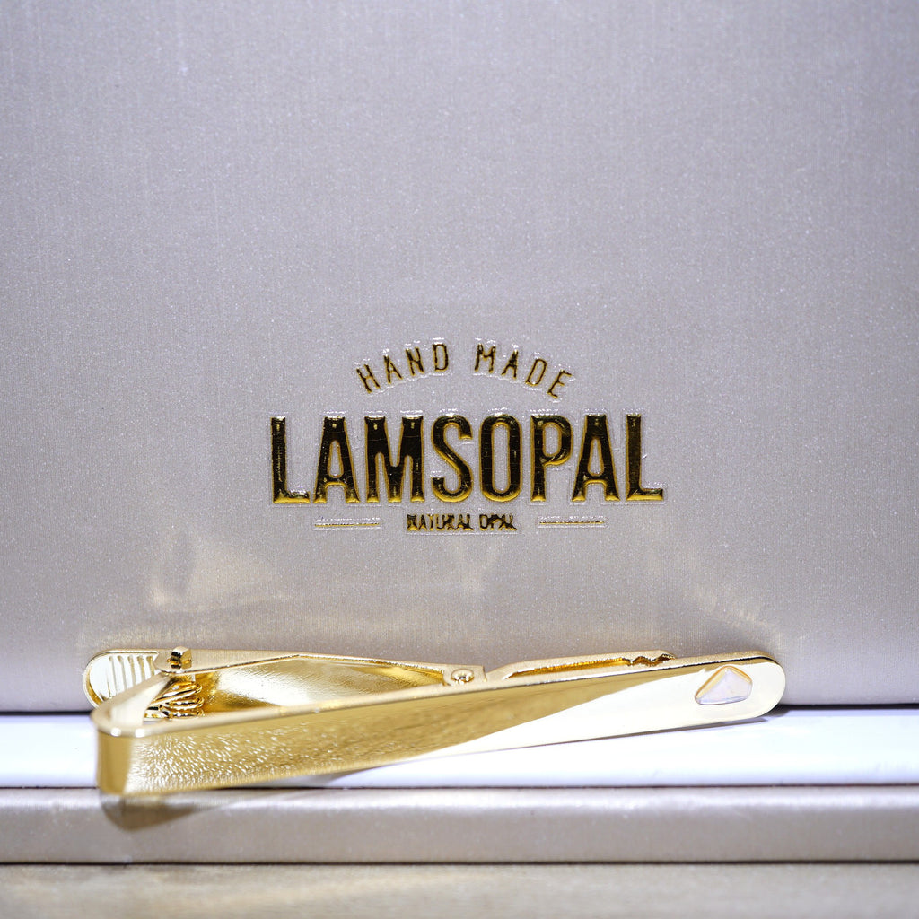 A gold tie bar with a textured finish and a polished clip, resting on a white surface in front of a silver-gray background with the brand name 'LAMSOPAL' printed in gold.