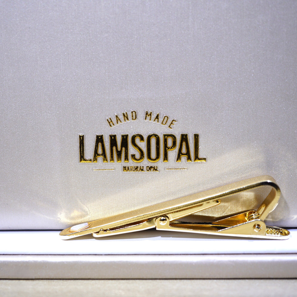 A gold tie bar clip resting on a white presentation box lid with the brand name 'LAMSOPAL' printed in gold foil.