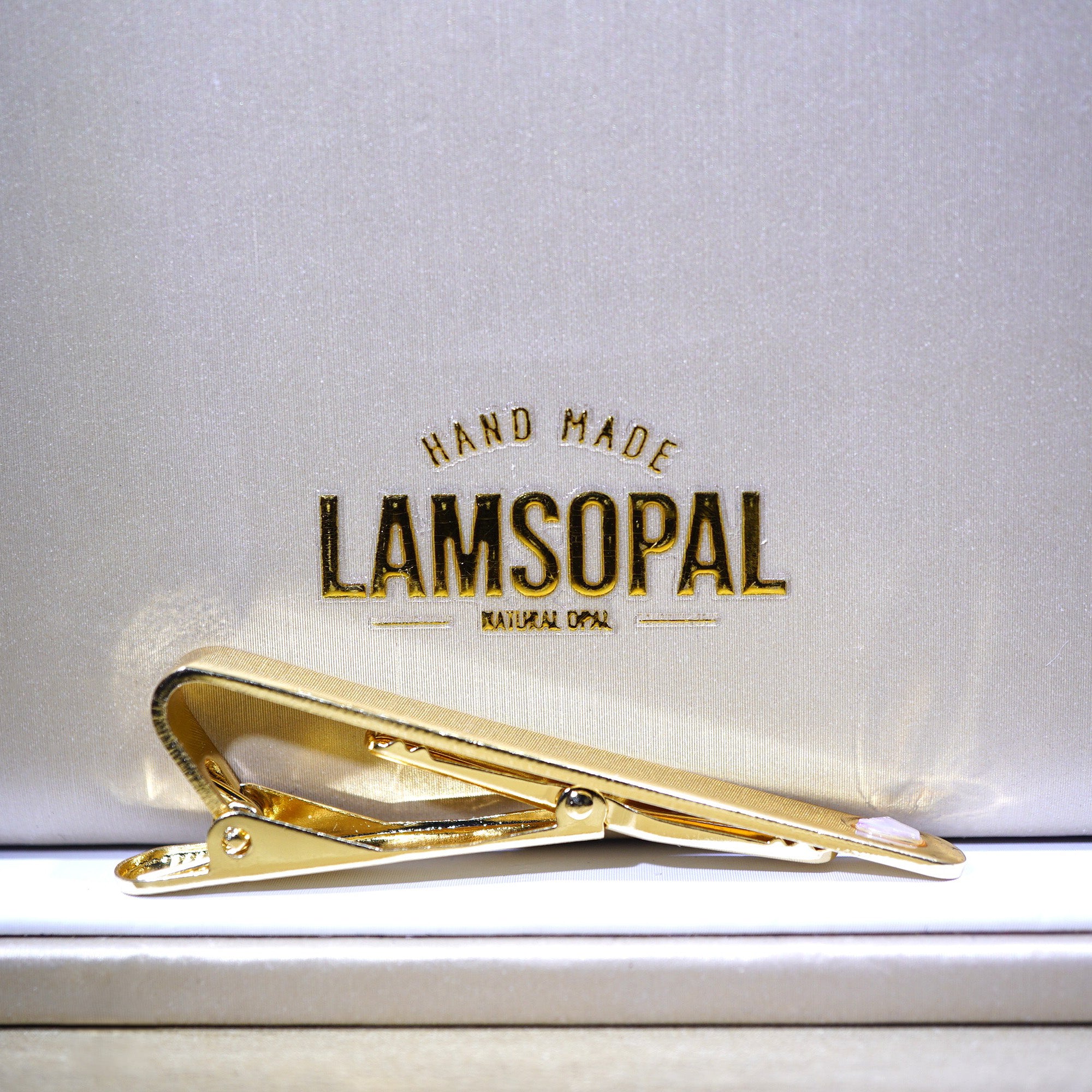 A gold-toned tie bar clip resting on a white surface in front of a branded Lamsopal box lid, with the text 'HAND MADE LAMSOPAL NATURAL OPAL' visible.