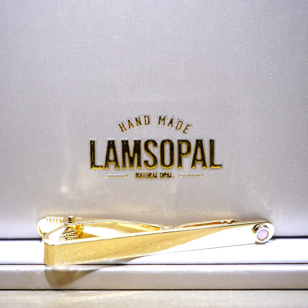 A gold Lamsopal tie clip resting on a metallic silver-gray presentation box lid, which features the embossed brand name 'LAMSOPAL' and 'HAND MADE' text.