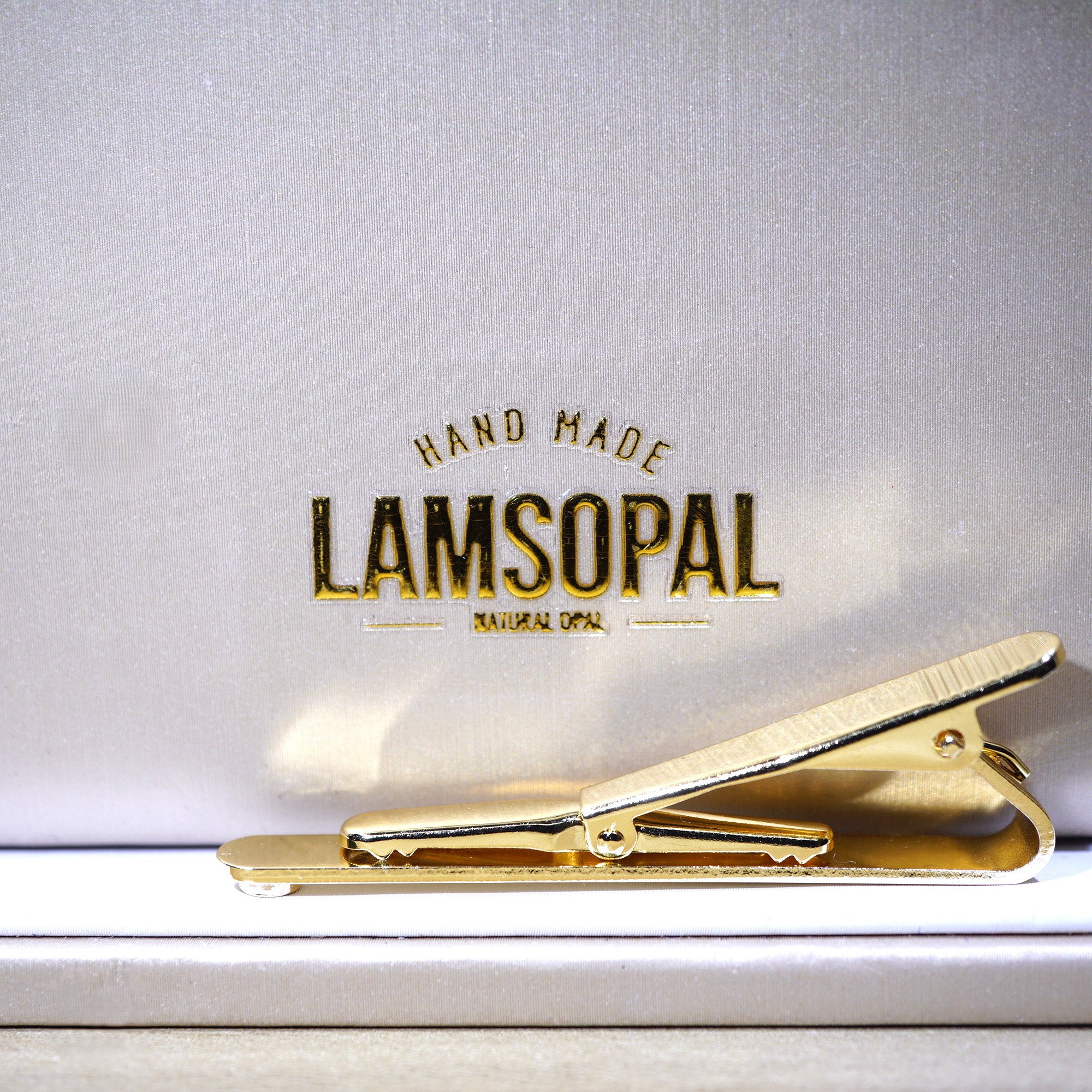 A gold metal hair clip resting on a white surface in front of a textured silver-gray background with the brand name 'LAMSOPAL' printed in gold.