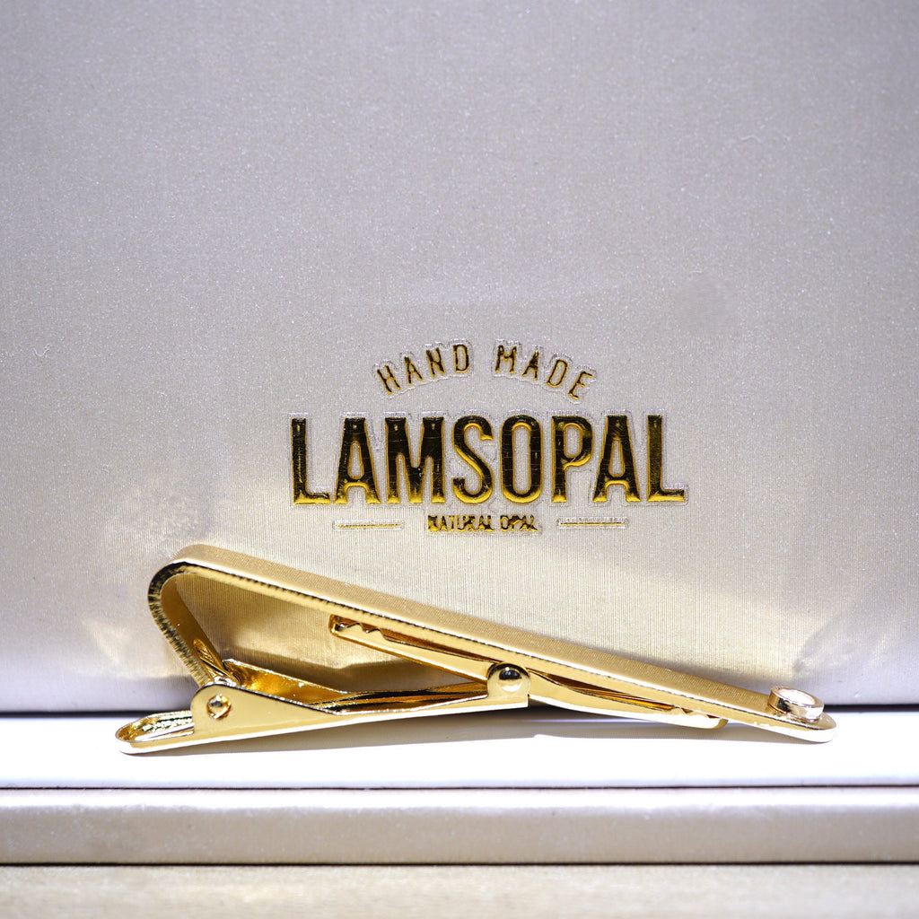 A shiny gold tie clip resting on a white textured surface with a branded backing card that reads 'HAND MADE LAMSOPAL'.