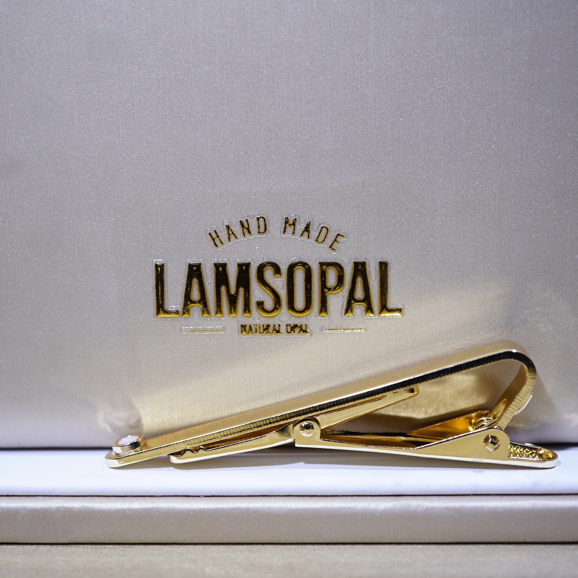 A gold tie bar clip resting on a white surface in front of a textured light gray background with the brand name 'LAMSOPAL' printed in gold.