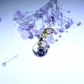 A silver-toned teardrop-shaped pendant with a dark blue iridescent stone in the center, hanging near dried light purple flowers on a pale background.