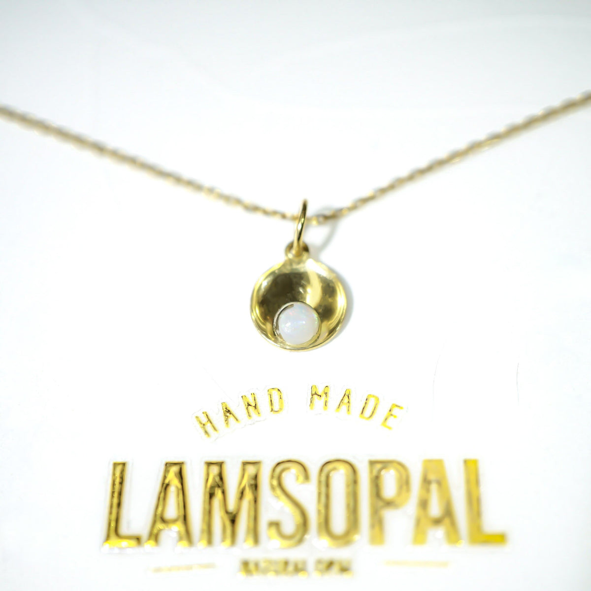 A gold necklace with a small teardrop-shaped pendant featuring a white inlay, displayed on a white background with the brand name 'LAMSOPAL' printed below.
