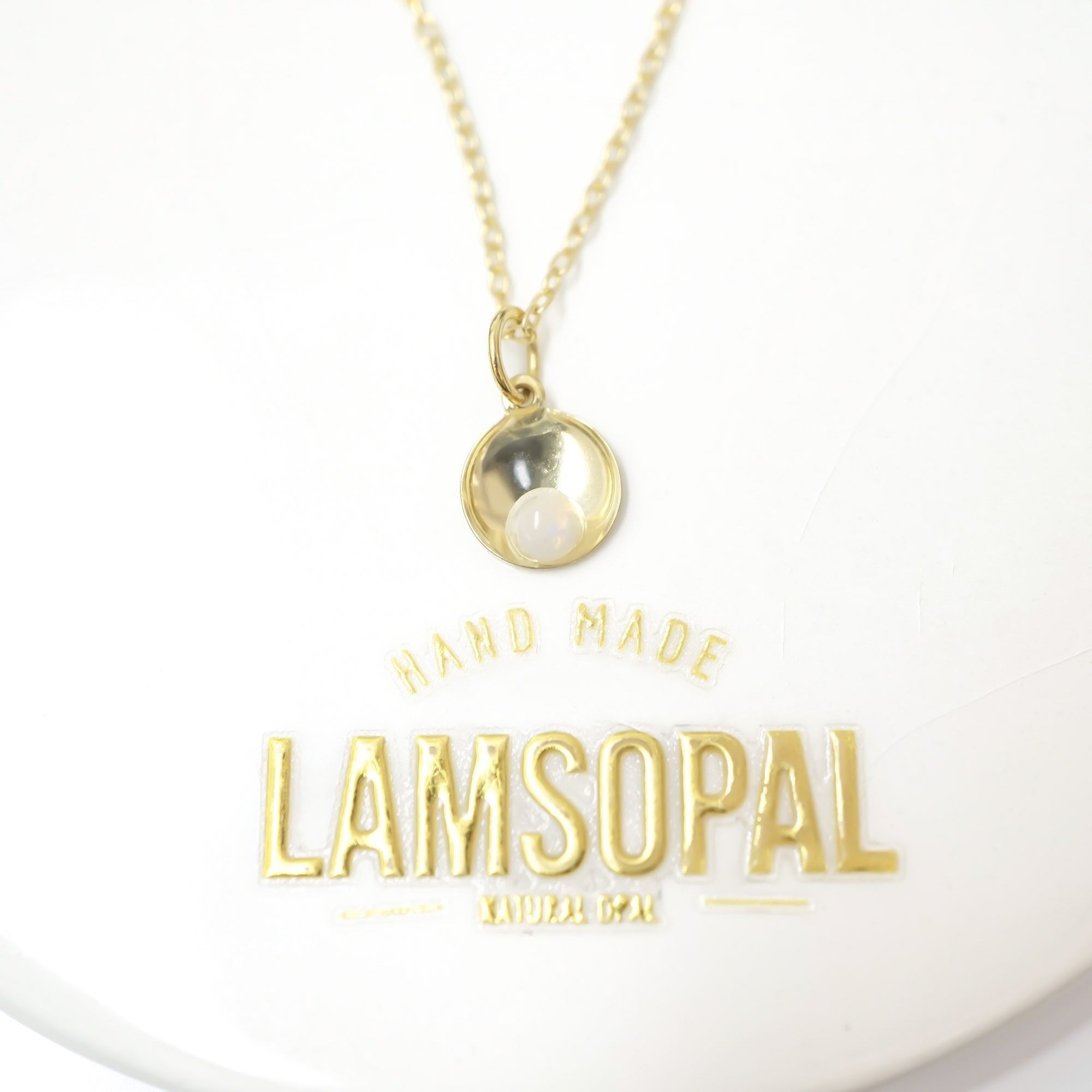 A gold necklace with a small oval pendant containing a white pearl, displayed on a white background with a gold 'LAMSOPAL HAND MADE NATURAL GEM' logo below.