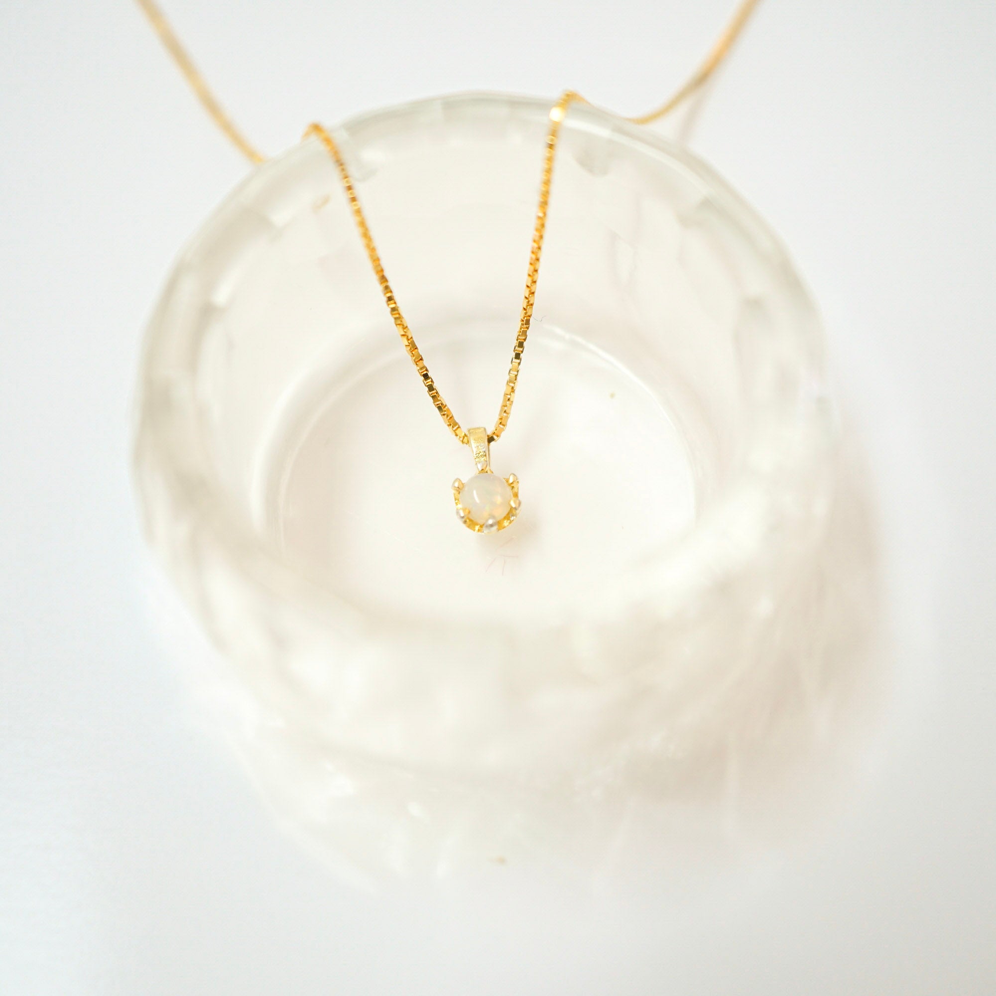 A delicate gold chain necklace with a small gold crescent moon pendant featuring a milky white stone, displayed on a textured clear glass dish against a white background.