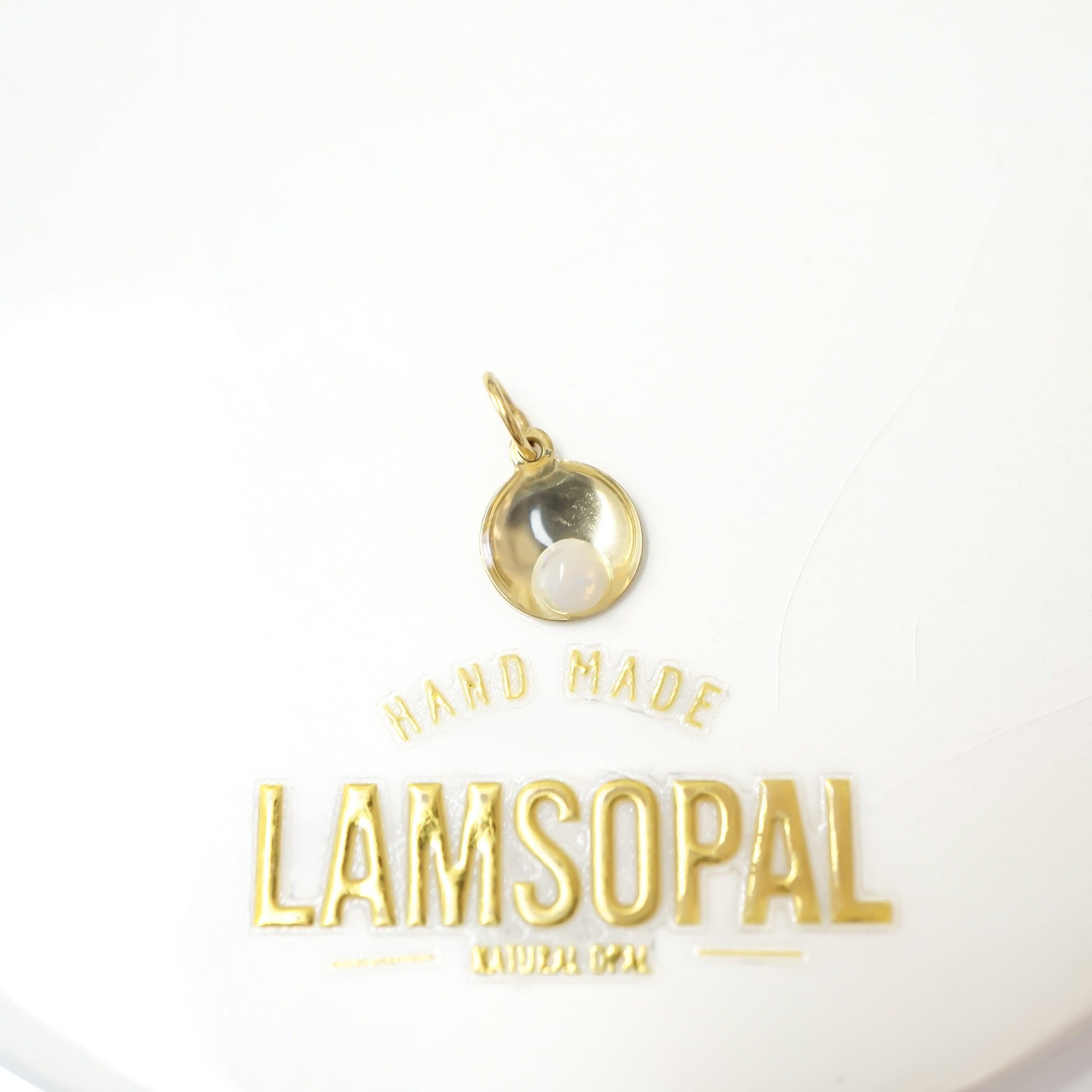 A small gold pendant charm with a white pearl-like center, displayed above gold text reading 'HAND MADE LAMSOPAL' on a white background.