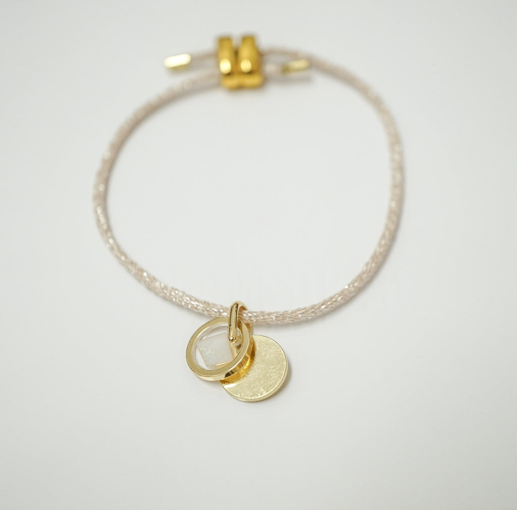 A delicate braided cord bracelet with a gold-toned adjustable clasp and two small hanging disc charms, one plain gold and one clear, shown on a light gray background.