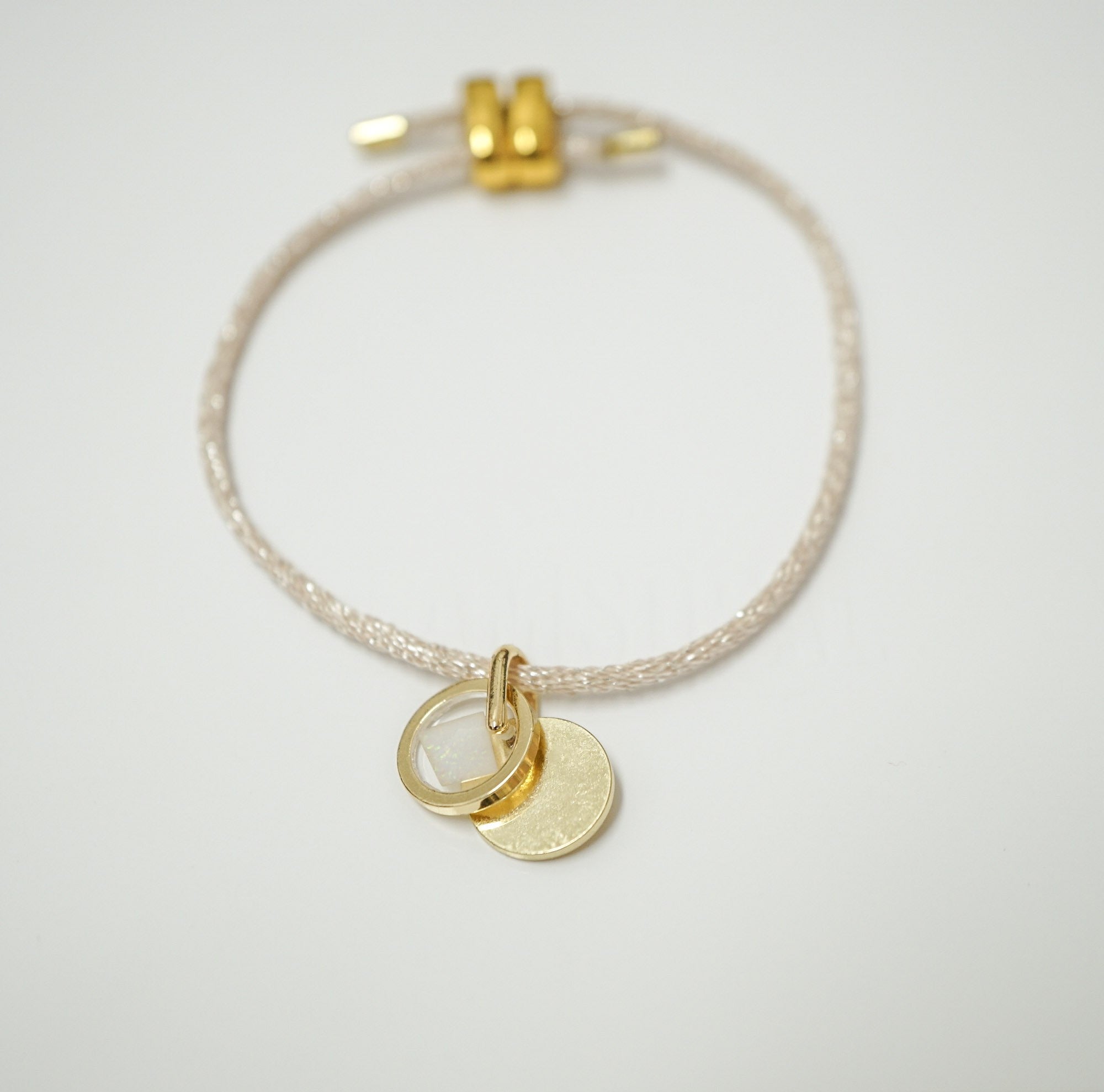 A delicate braided cord bracelet with a gold-toned adjustable clasp and two small hanging disc charms, one plain gold and one clear, shown on a light gray background.