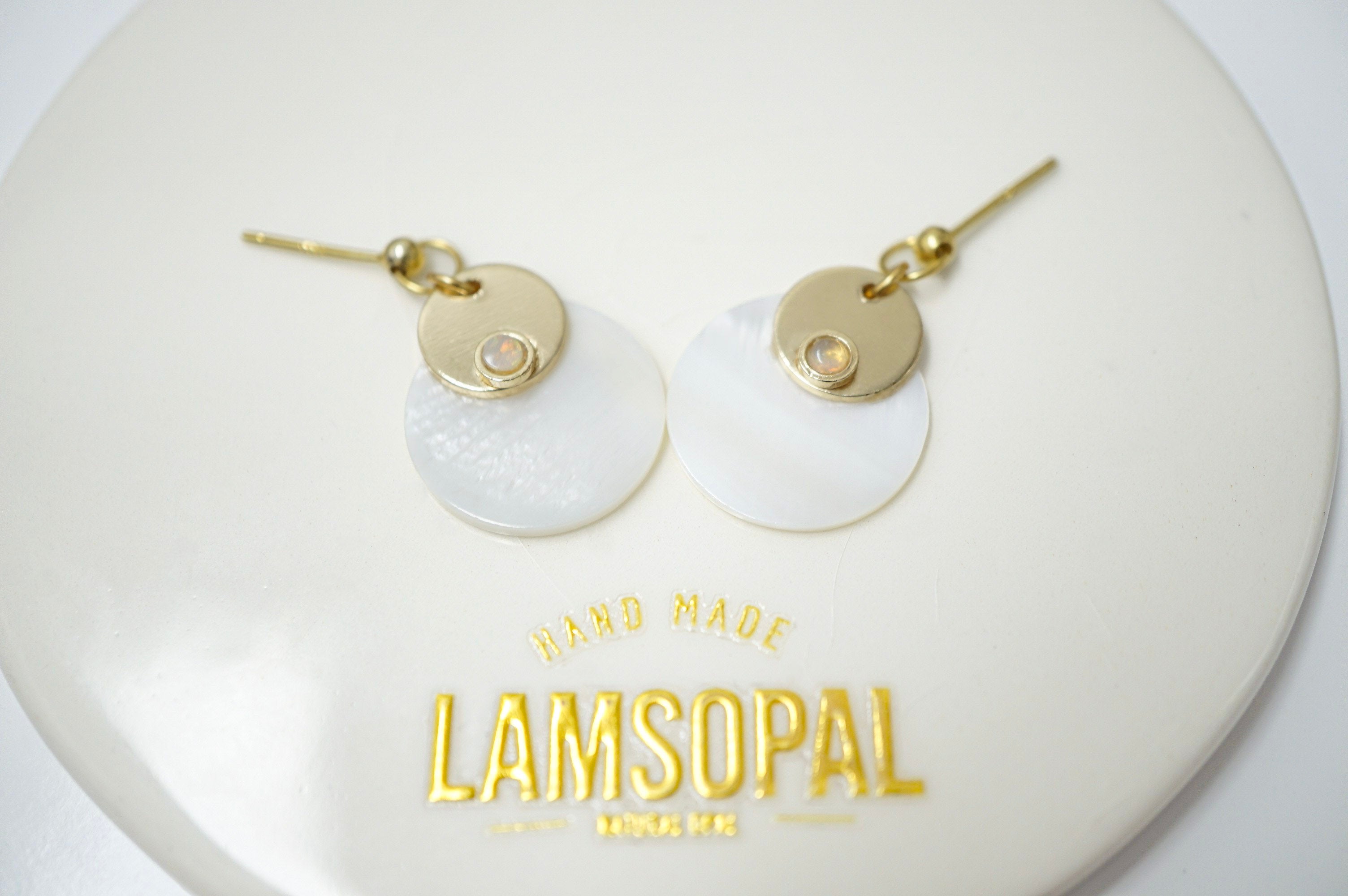 A pair of gold and white disc drop earrings displayed on a white circular stand with the brand name 'LAMSOPAL' printed in gold below.