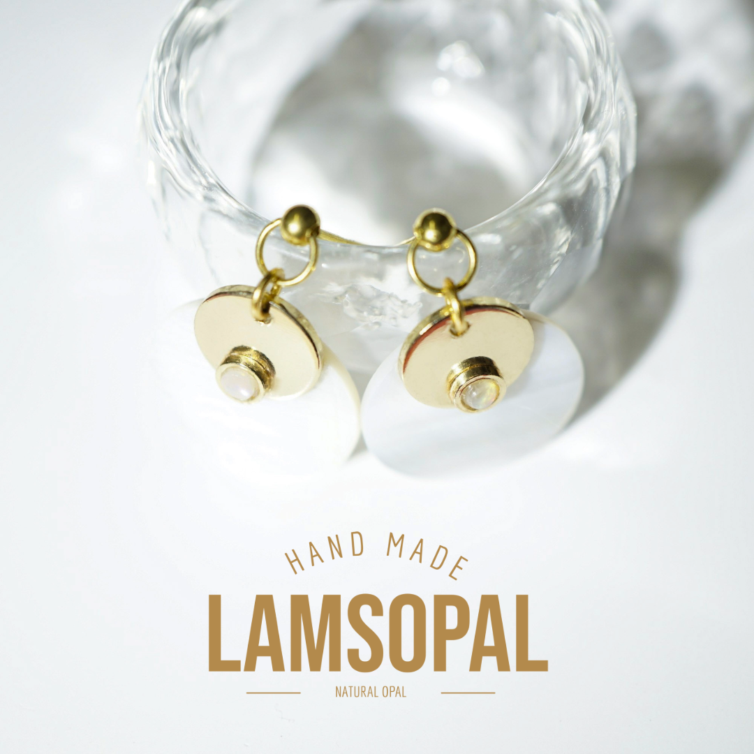 A pair of handmade Lamsopal natural opal dangle earrings with gold-plated circular discs and small pearl-like center accents, hanging from a clear glass tumbler. The text 'HAND MADE LAMSOPAL NATURAL OPAL' is displayed below.