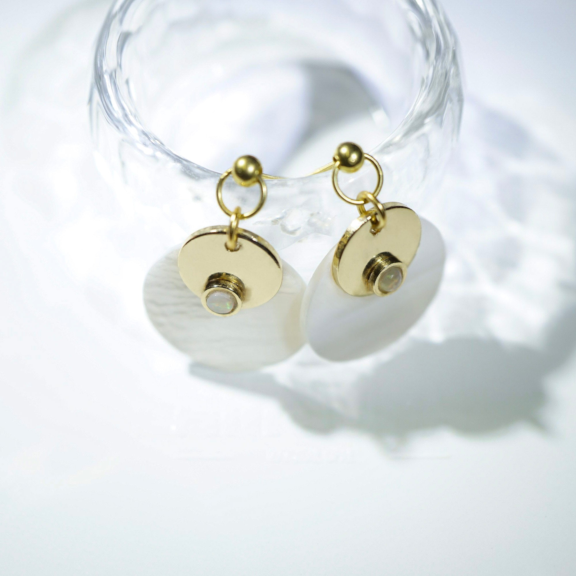 A pair of gold-tone dangle earrings with round disc charms and small white inlay details, displayed on a clear glass surface against a bright white background.