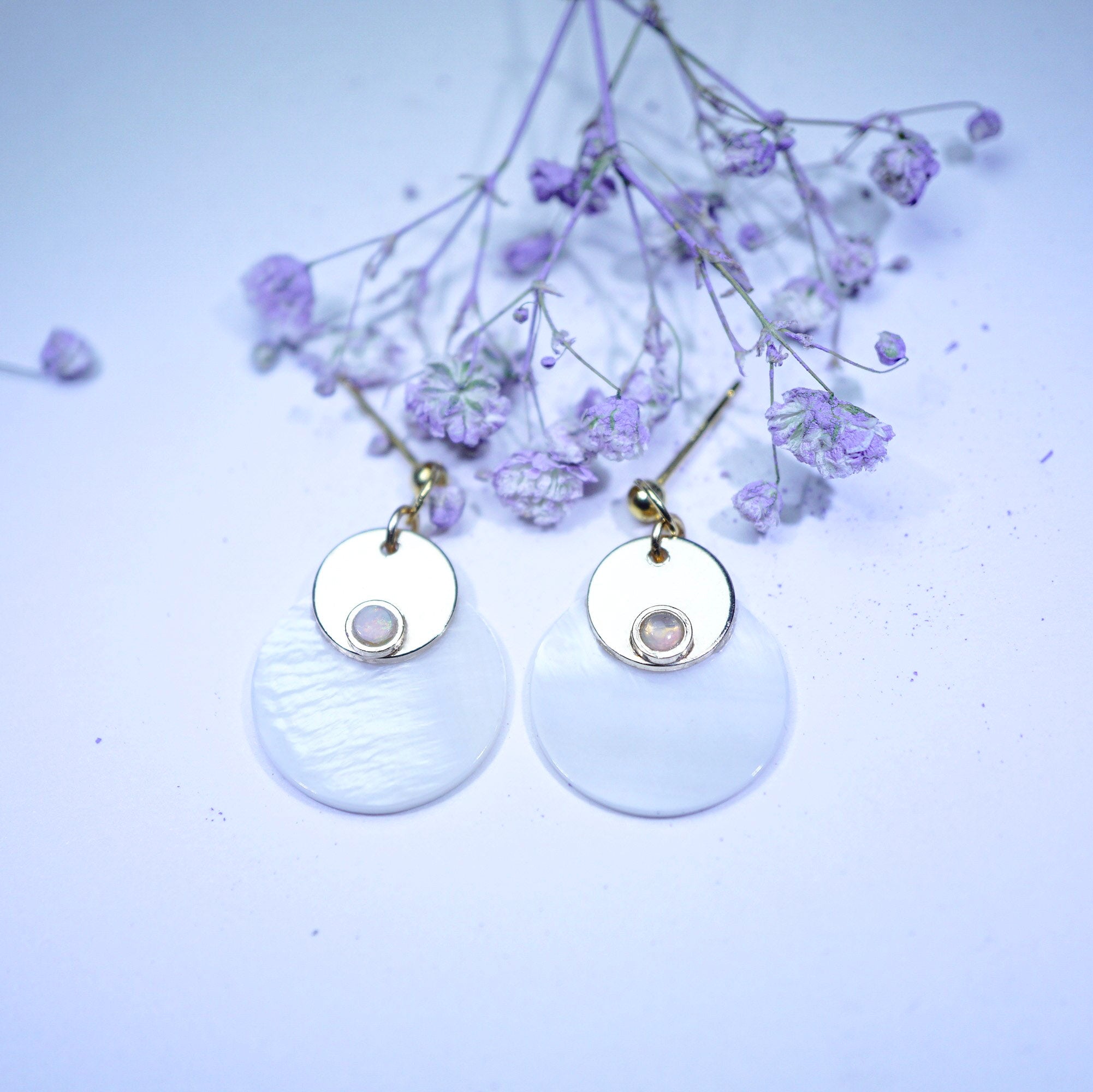 A pair of dangle earrings with gold-tone metal and white circular discs, displayed on a light lavender background with dried purple flowers.