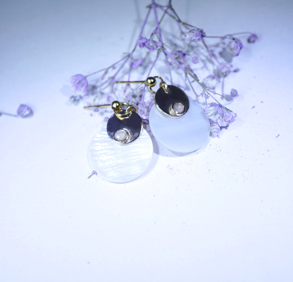 A pair of dangle earrings with gold-toned posts, dark round metal discs, and large milky white oval pendants, styled with small dried lavender flowers on a pale background.
