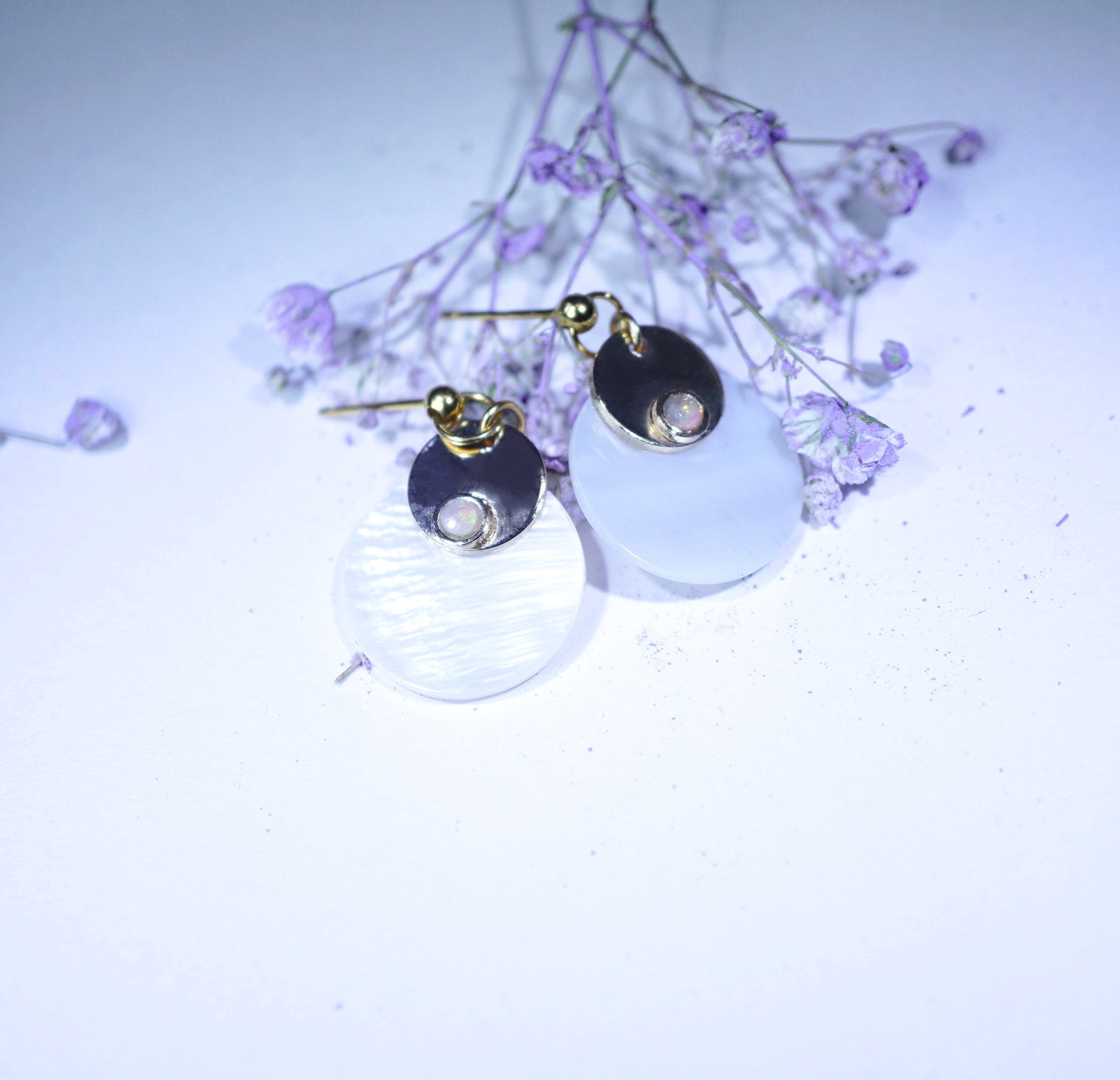 A pair of dangle earrings with gold-toned posts, dark round metal discs, and large milky white oval pendants, styled with small dried lavender flowers on a pale background.