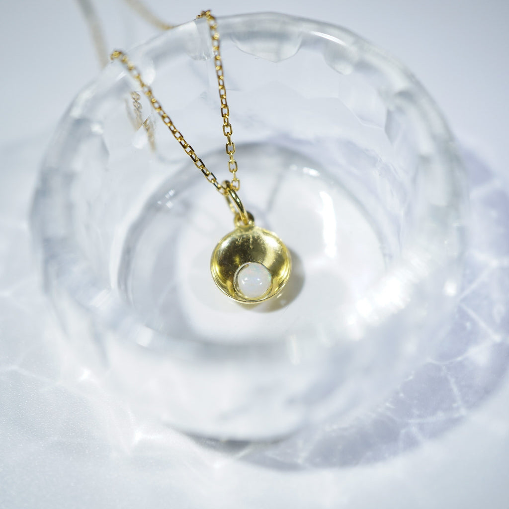 A gold pendant necklace with a small, textured gold dome and a white inlay, resting inside a clear glass dish on a white surface.