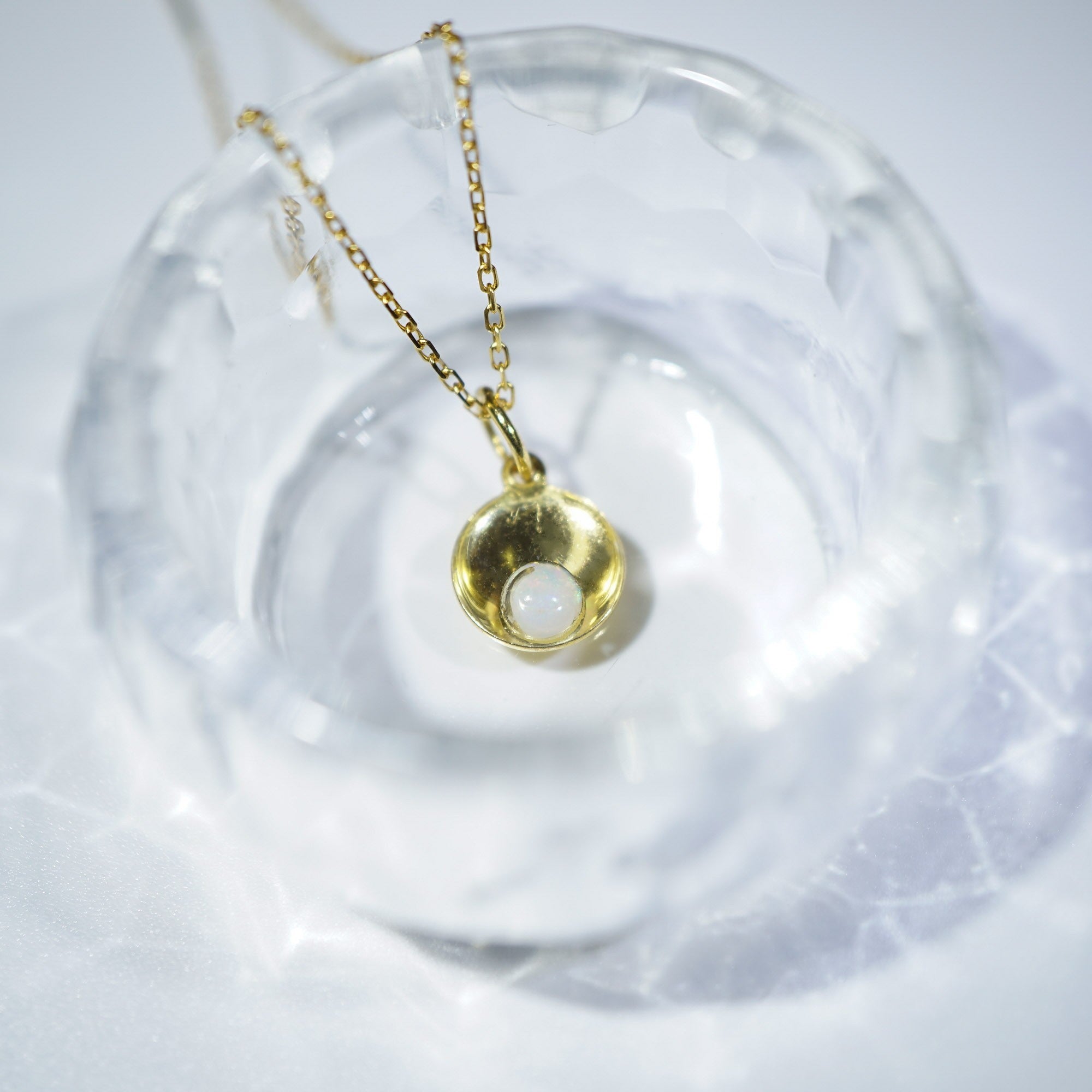 A gold pendant necklace with a small, textured gold dome and a white inlay, resting inside a clear glass dish on a white surface.