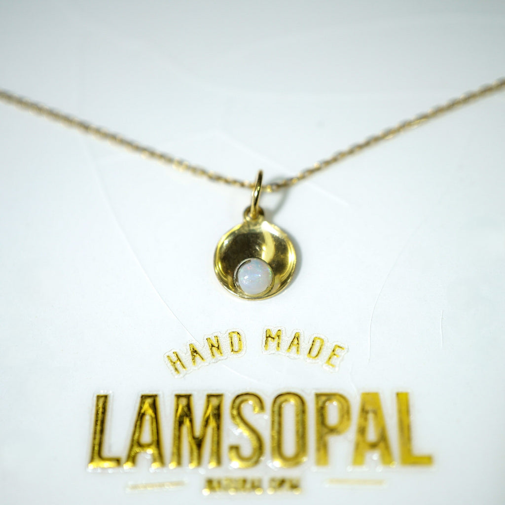 A gold necklace with a small crescent-shaped pendant and a white stone, displayed on a white card with the brand name 'LAMSOPAL' and 'HAND MADE' printed in gold.