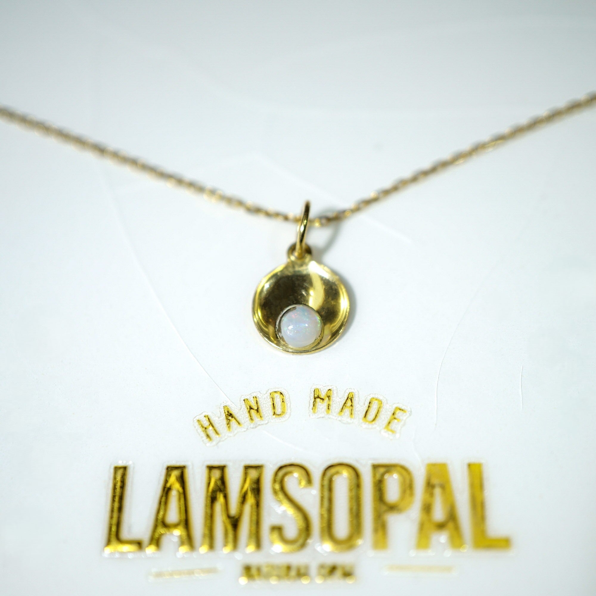 A gold necklace with a small crescent-shaped pendant and a white stone, displayed on a white card with the brand name 'LAMSOPAL' and 'HAND MADE' printed in gold.