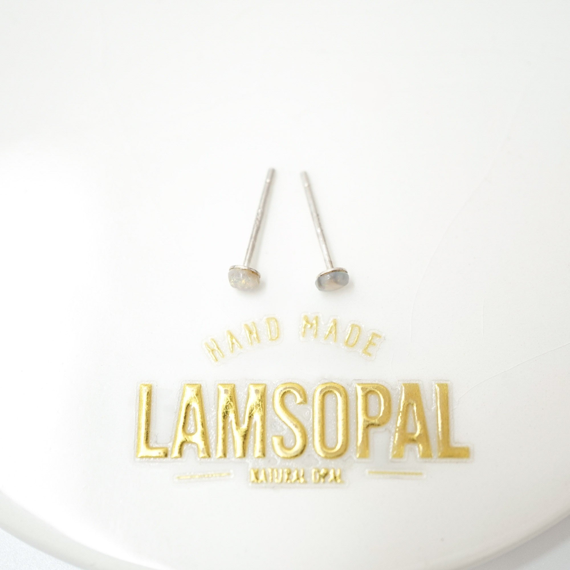 Two small silver stud earrings with flat oval gray-blue tops are displayed on a white surface above gold foil branding that reads 'HAND MADE LAMSOPAL NATURAL GAN'.