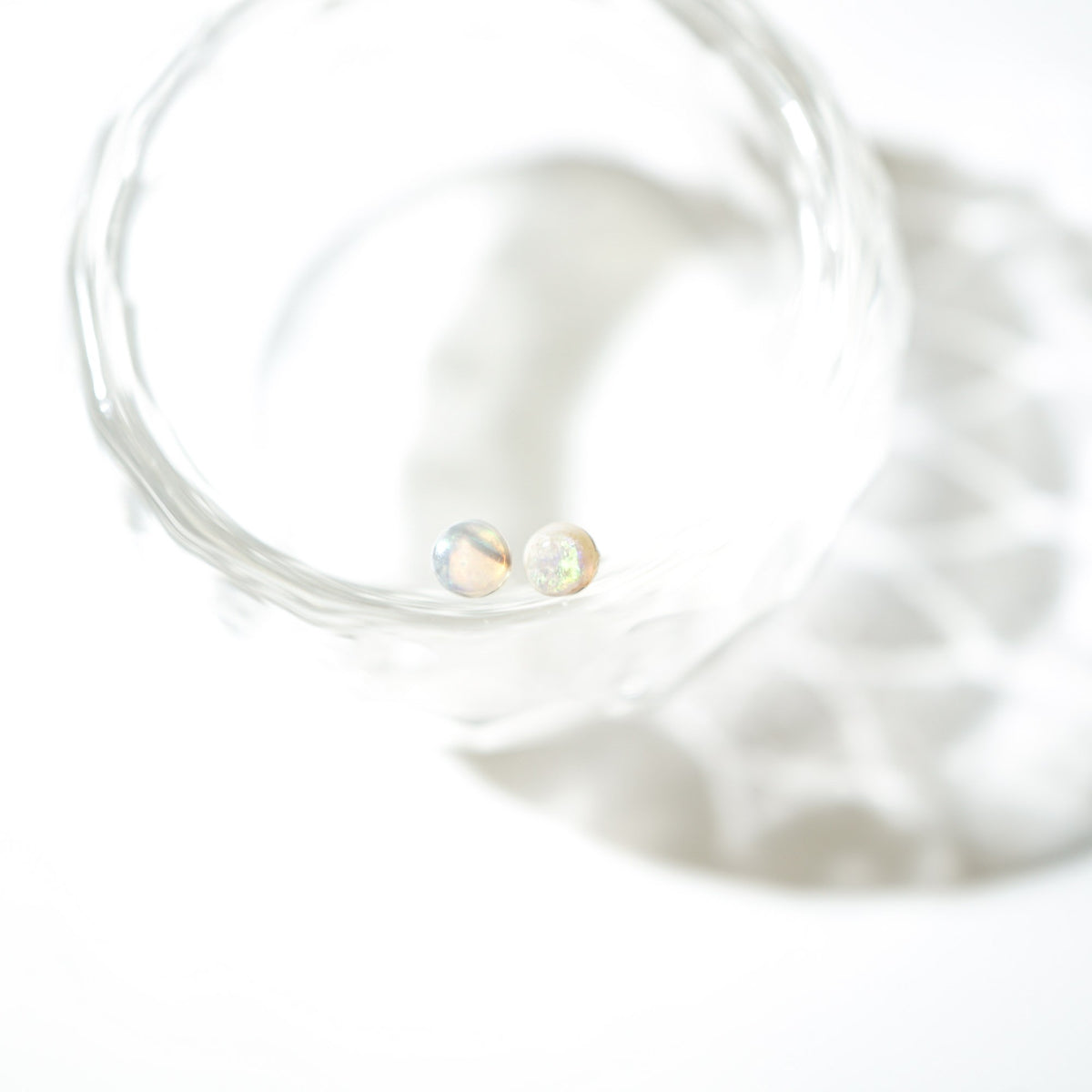 Two small round iridescent cabochon beads resting inside a clear glass dish on a bright white background.