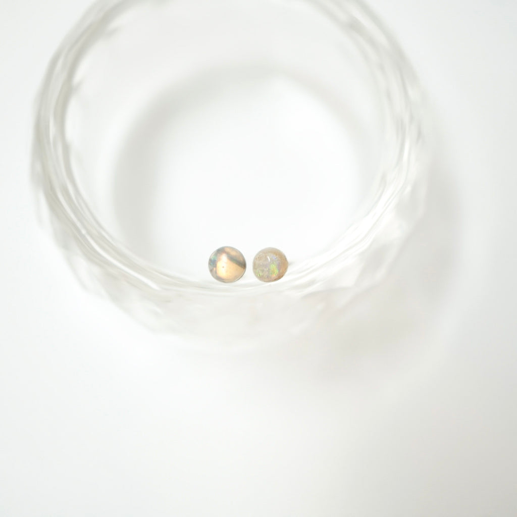 A pair of small round stud earrings with iridescent, opalescent surfaces resting inside a clear textured glass bowl on a white background.