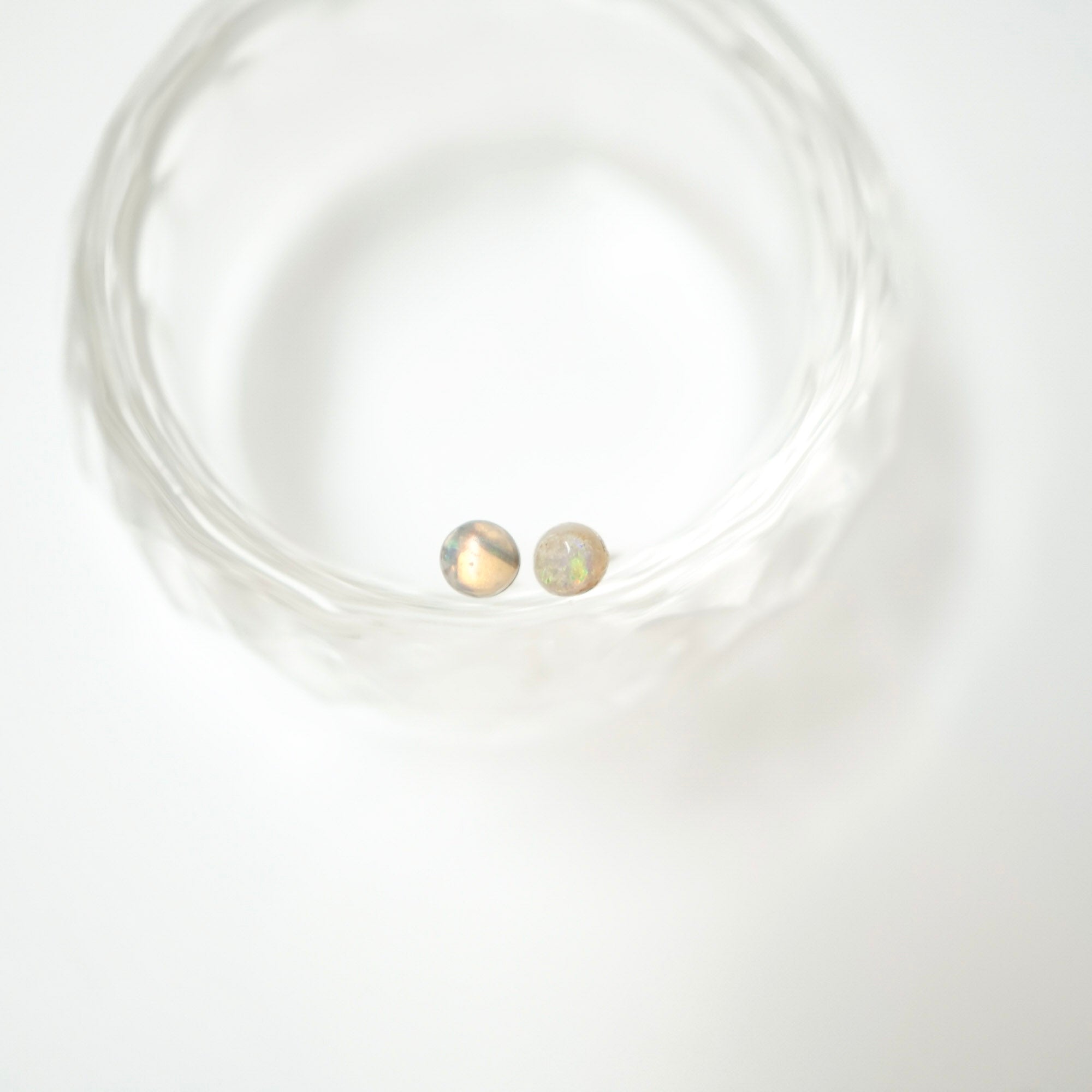 A pair of small round stud earrings with iridescent, opalescent surfaces resting inside a clear textured glass bowl on a white background.