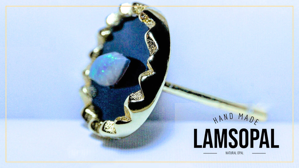 A handmade Lamsopal stud earring featuring a natural opal in a bezel setting with black enamel and gold accents, shown on a light blue background with a brand logo overlay.