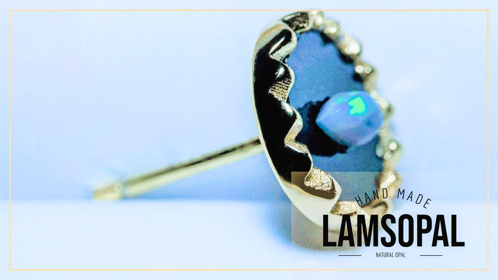 A gold-toned Lamsopal stud earring with a black bezel-set natural opal showing blue and green flashes, displayed on a light blue background with a 'LAMSOPAL' brand overlay.