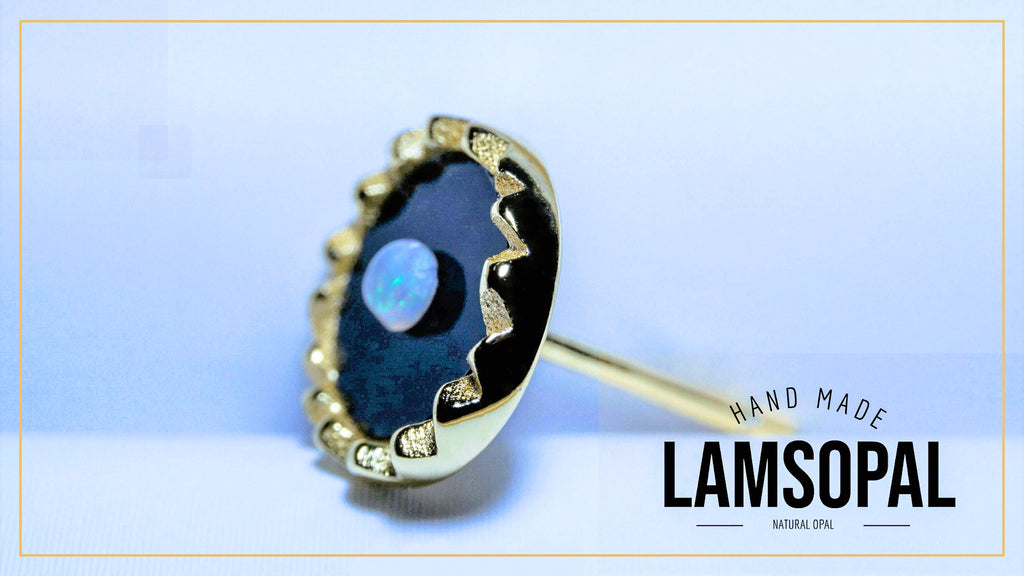 A single Lamsopal stud earring featuring a natural opal inlay with a blue iridescent center surrounded by a black and gold scalloped bezel setting. The brand name 'LAMSOPAL' is visible in the bottom right corner.
