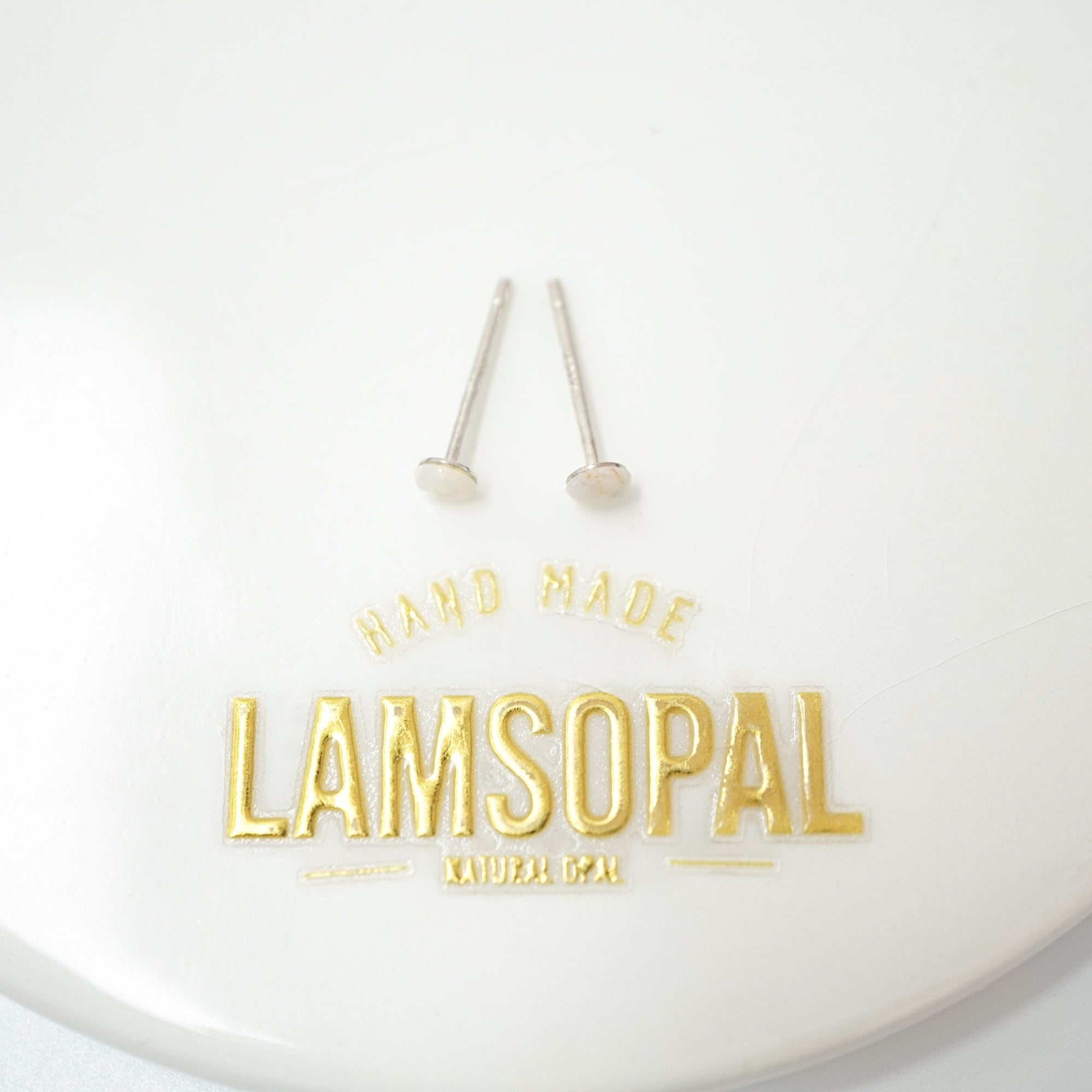 Two small gold post earrings displayed above a white oval surface with gold text reading 'HAND MADE LAMSOPAL NATURAL GEM'.