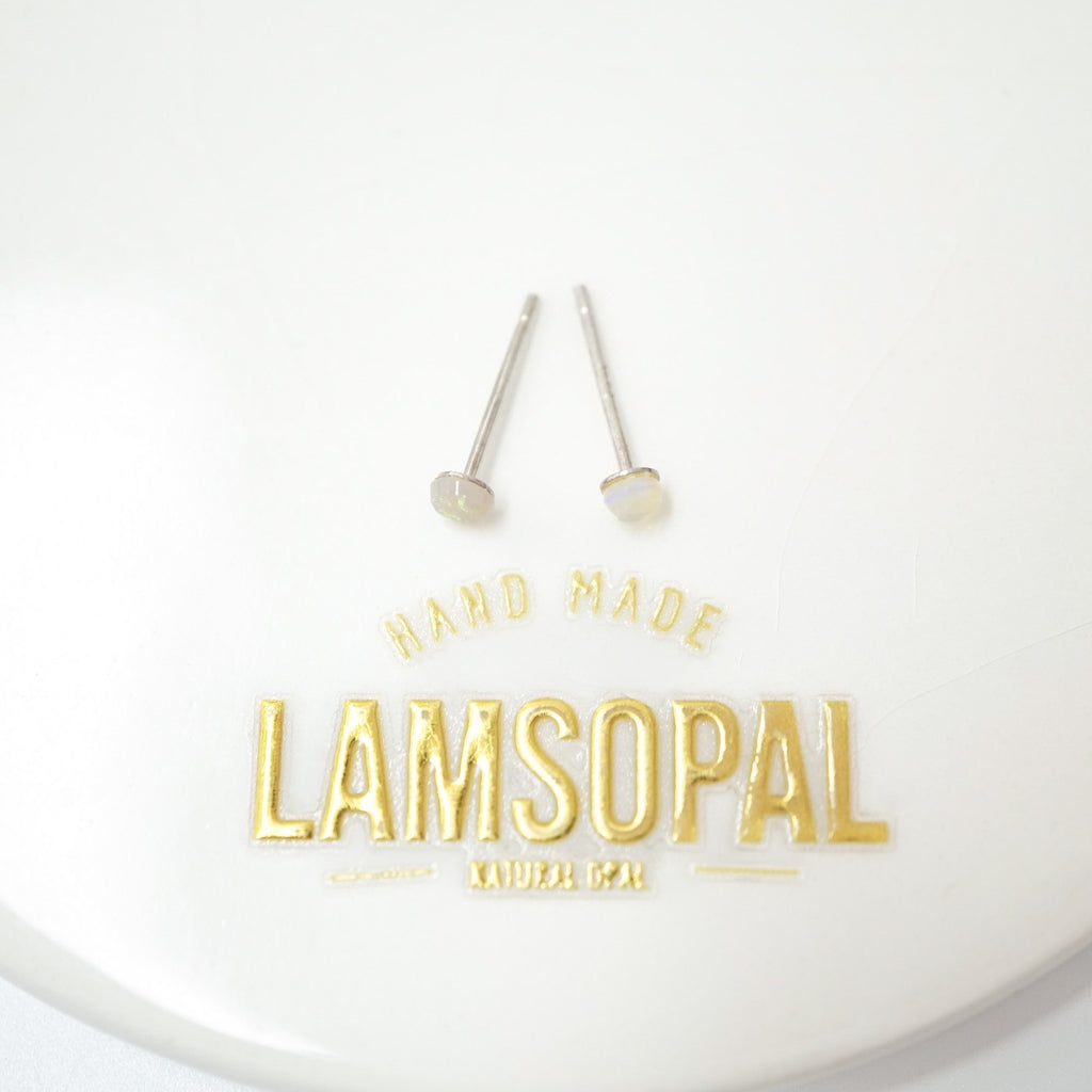 A pair of silver-toned stud earrings with round white stones, displayed on a white surface above gold foil branding that reads 'HAND MADE LAMSOPAL'.