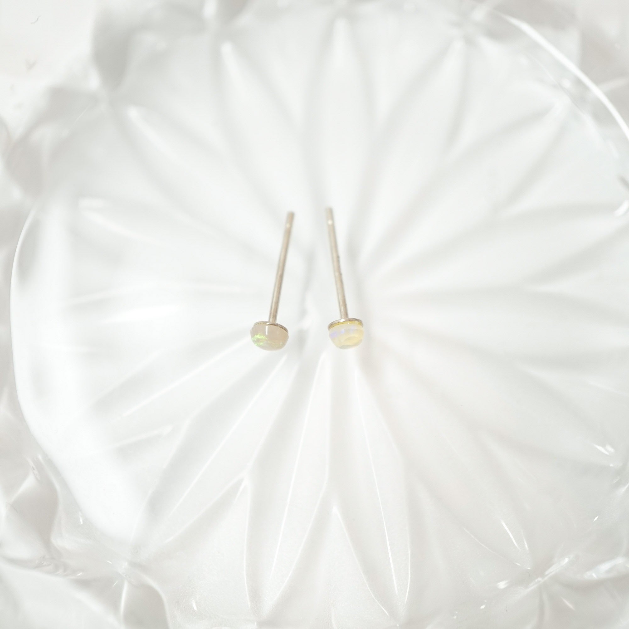 A pair of small gold stud earrings with oval iridescent opal-like beads, displayed on a textured white glass plate.