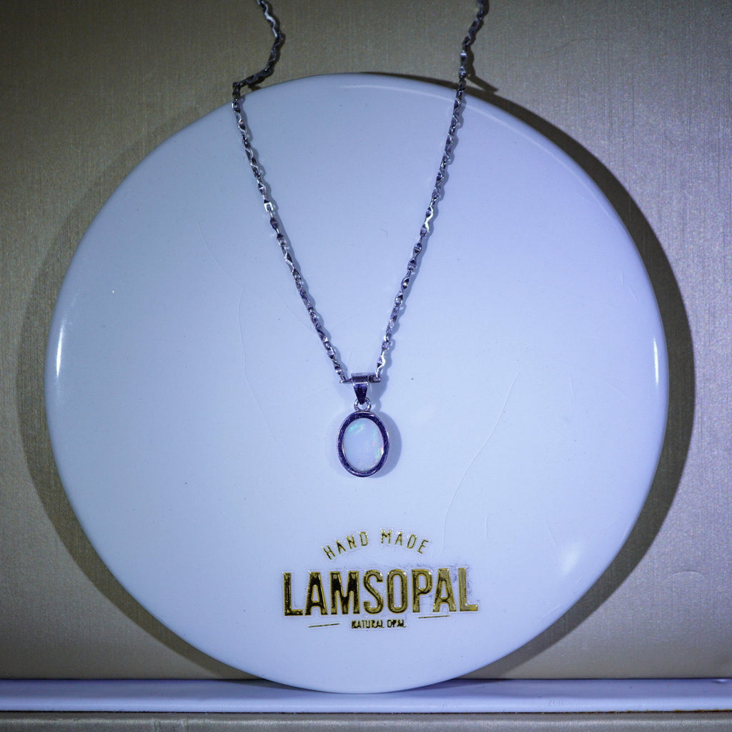 A silver-toned chain necklace with a simple oval pendant displayed on a round white branded stand. The stand has the brand name 'LAMSOPAL' printed in gold near the bottom.