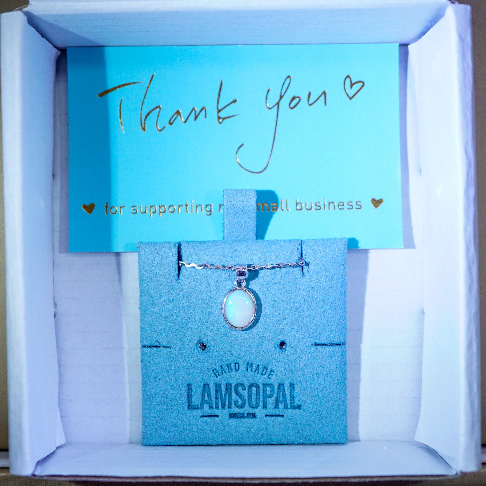 A silver-toned necklace with a round iridescent pendant displayed on a blue card inside a white gift box. The box contains a blue card with the brand name LAMSOPAL and a handwritten 'Thank you' note.