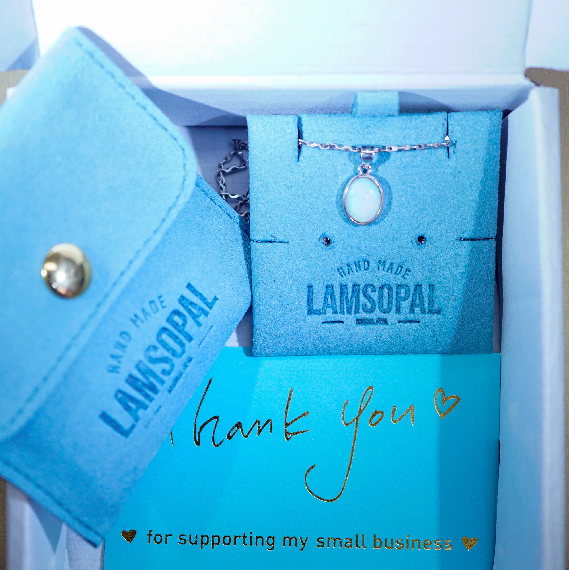 A Lamsopal necklace with a round white stone pendant displayed in its branded light blue gift box, alongside a matching blue pouch and thank-you card.