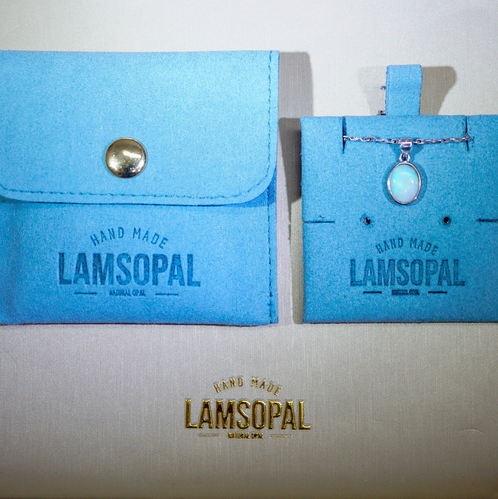 A light blue fabric jewelry pouch with a snap closure and a matching display card holding a small iridescent pendant necklace, both branded Lamsopal, on a light beige background.